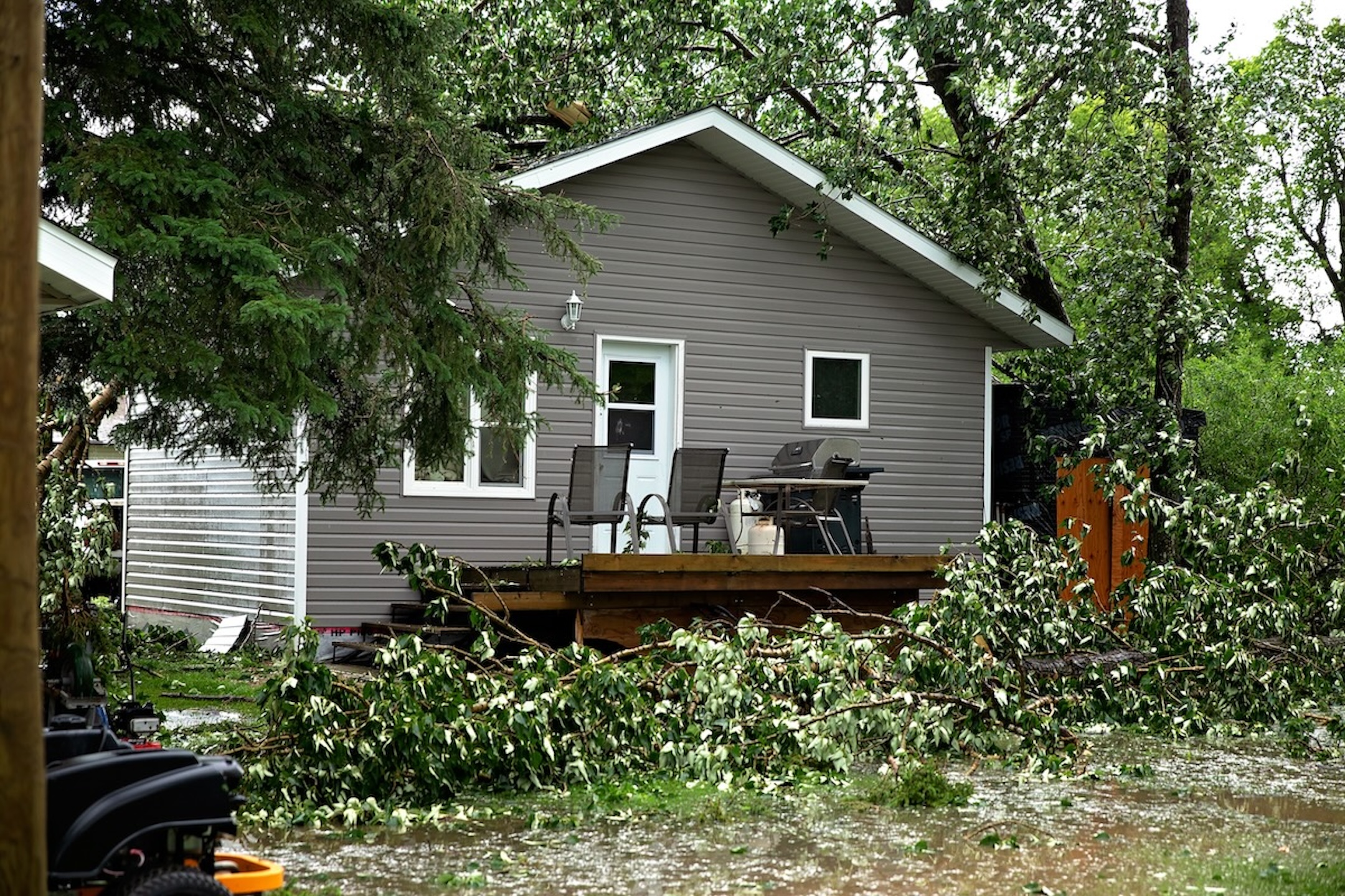 A grey home with white trim and a wooden deck surrounded by fallen tree limbs and debris, highlighting the immediate need for a storm damage inspection to check for structural roof issues.