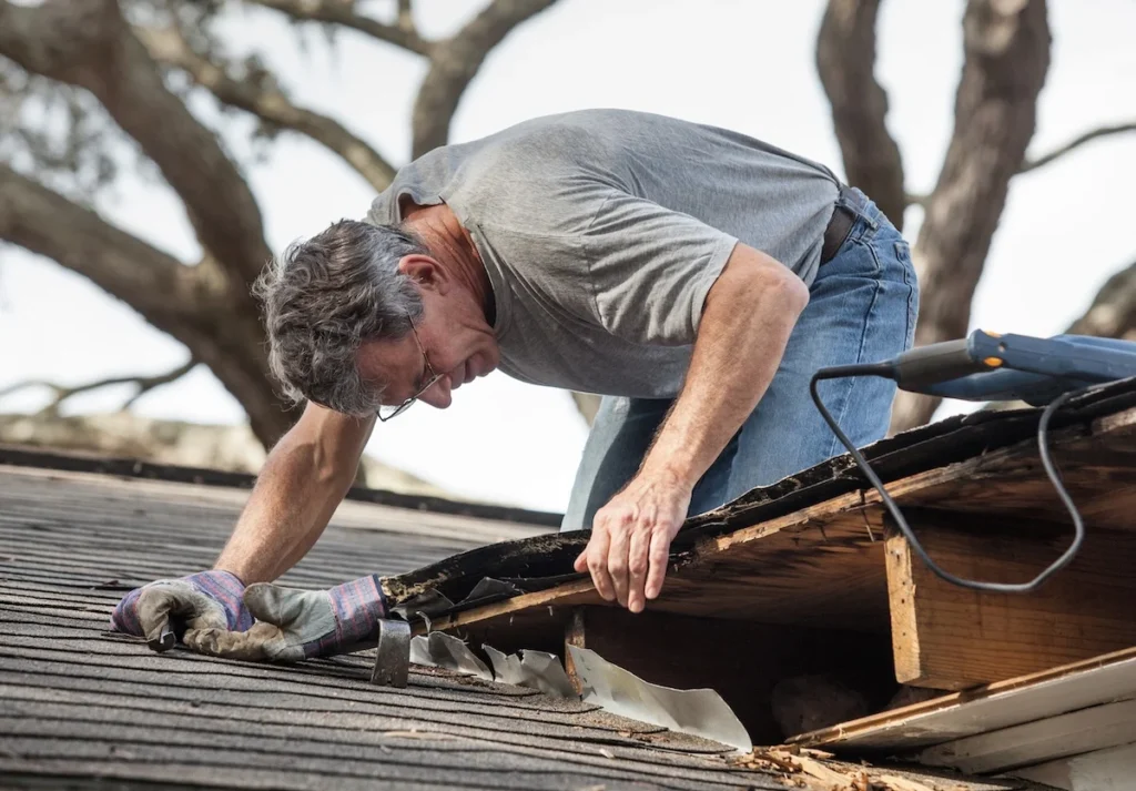 A roofing contractor working to tear out and assess severe damage near the edge of a shingle roof.