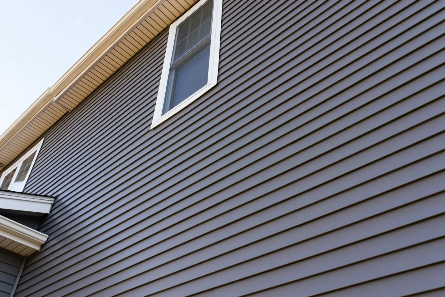 A detailed architectural view of premium dark grey horizontal vinyl siding installed on a residential property, featuring a white-trimmed window and a clean tan soffit system.