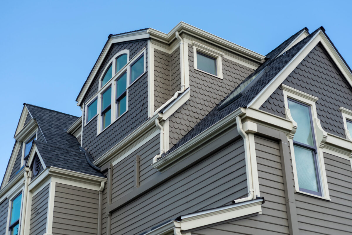 A multi-story large historic home under blue sky featuring intricate scalloped shake siding, multiple gabled rooflines with charcoal shingles, and a sophisticated white gutter.